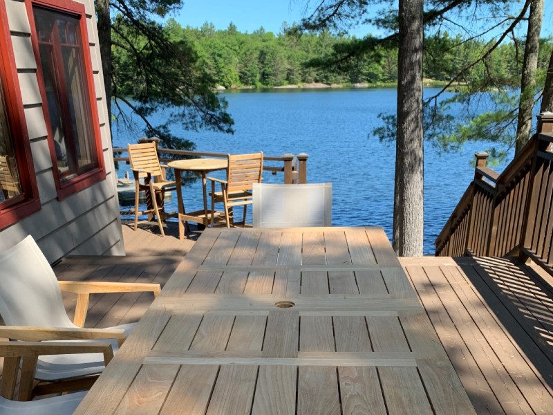 Light teak rectangular table with white sling arm chairs in front of a lake